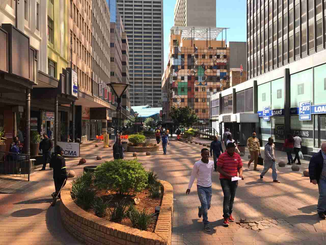 Pedestrian-friendly city street with people walking among landscaped planters and modern building facades.