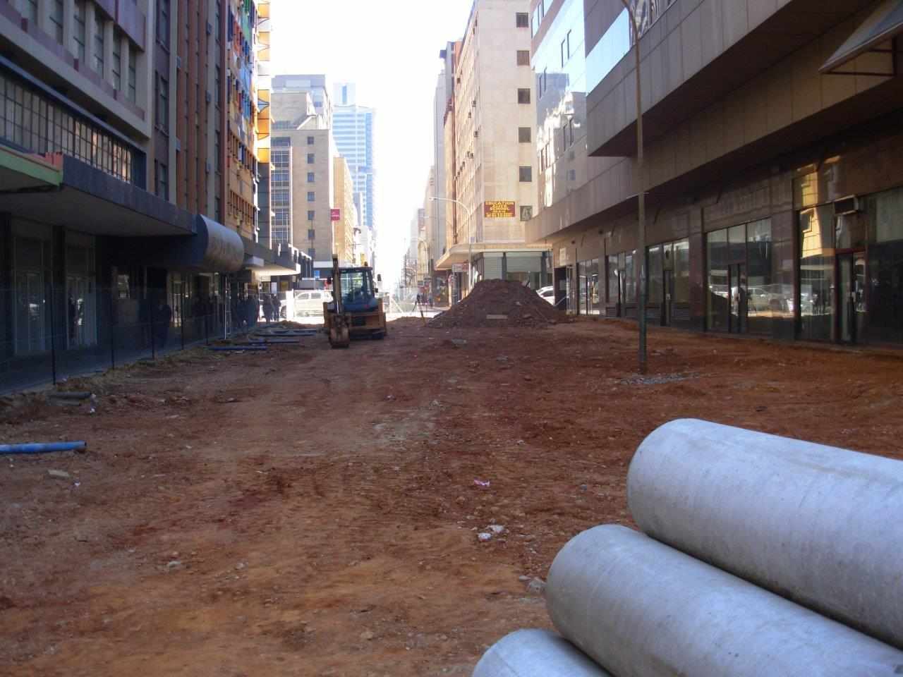 Street construction site with heavy machinery leveling soil between tall commercial buildings.
