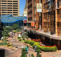 Pedestrian walkway in the Johannesburg CBD with landscaped gardens, people walking, and tall office buildings in the background.