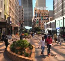 Urban pedestrian walkway with landscaped planters, shops, and people walking through the city.