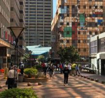Pedestrian walkway in a busy city center with people walking between landscaped planters and tall buildings.